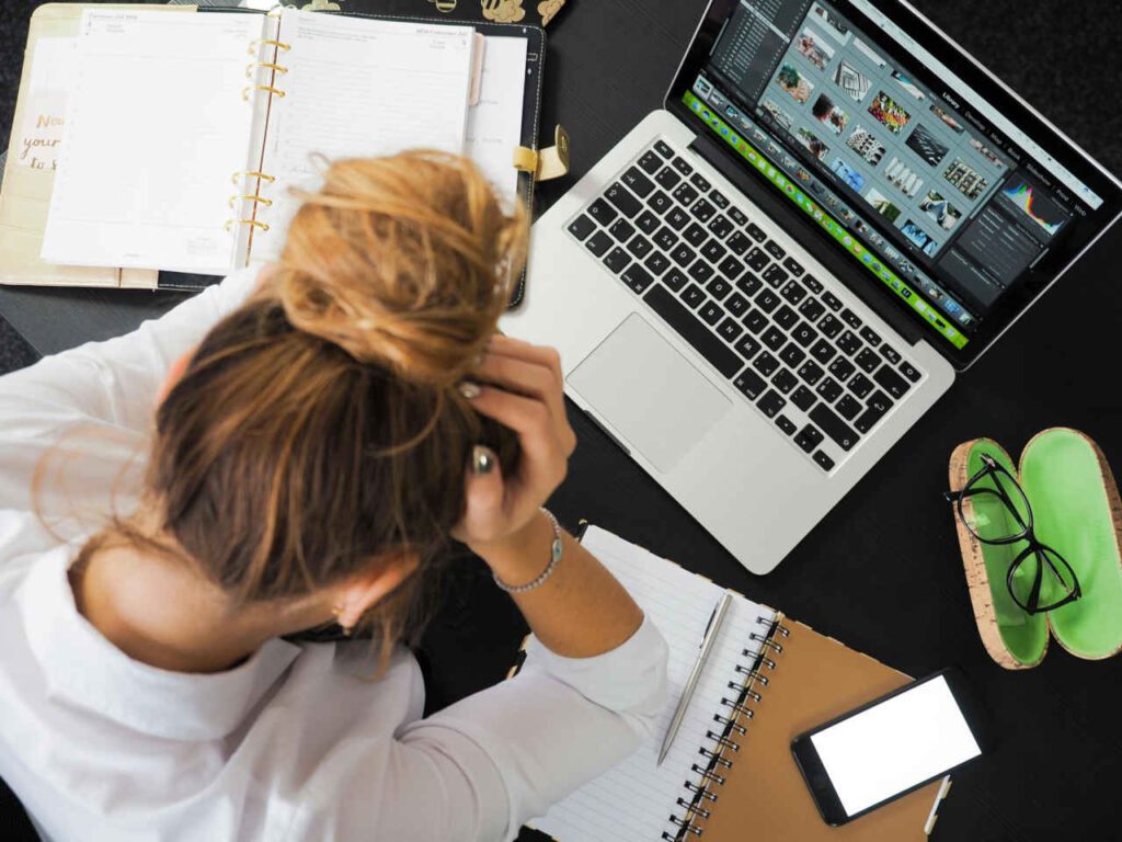 Persona estresada trabajando en un escritorio con una computadora portátil, cuaderno, agenda, teléfono móvil y gafas