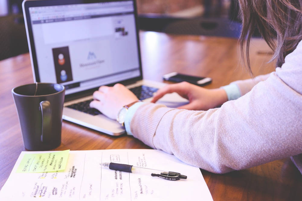 Persona escribiendo en una computadora portátil en un escritorio con una taza, bolígrafo y notas manuscritas