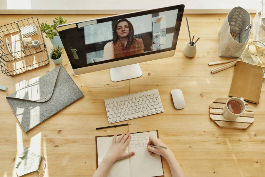 Persona tomando notas durante un curso en línea en videollamada, con escritorio ordenado y artículos de oficina