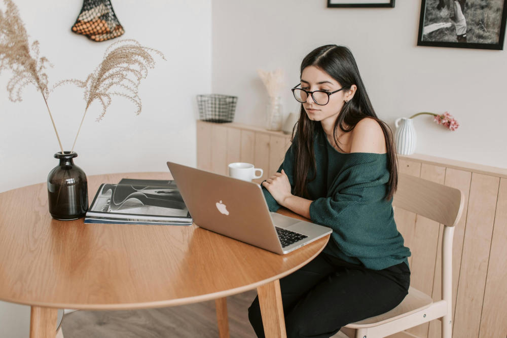 Persona con gafas trabajando en una computadora portátil en una mesa redonda de madera con un jarrón con ramas decorativas, una taza y algunos libros