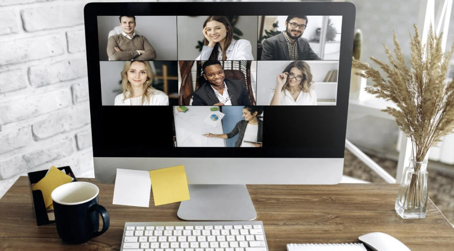 Videoconferencia con seis participantes en una computadora de escritorio, sobre un escritorio con taza de café, notas adhesivas y jarrón con ramas decorativas